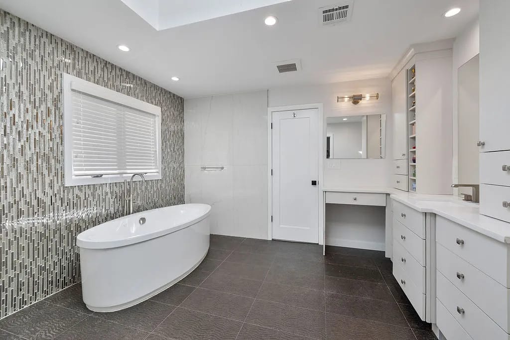 Bathroom with freestanding oval bathtub, dual sink vanity, white cabinetry, and mosaic tiled accent wall.