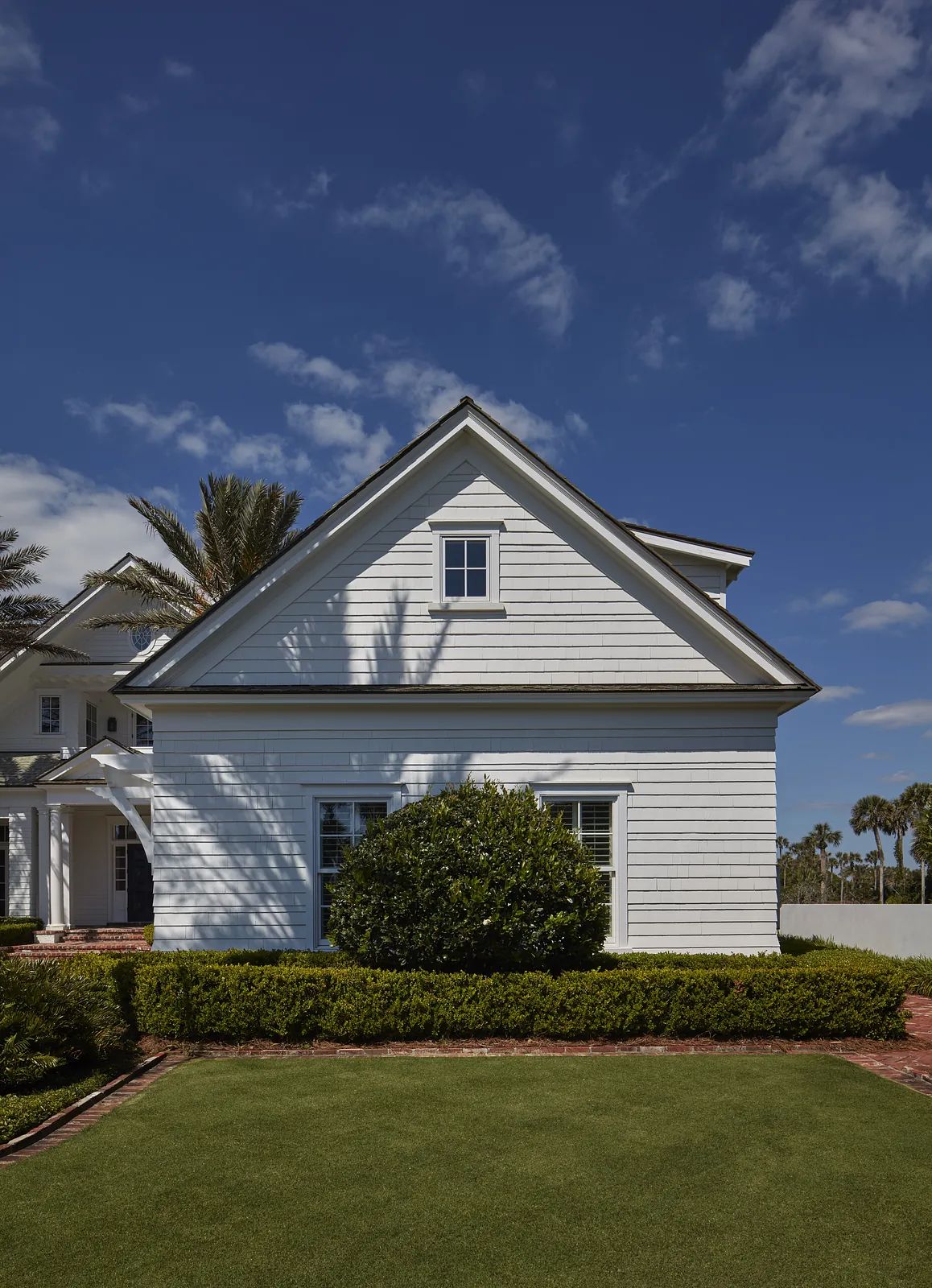 Exterior of a house with white siding, gable roof, window, bush, lawn, and palm trees against a blue sky
