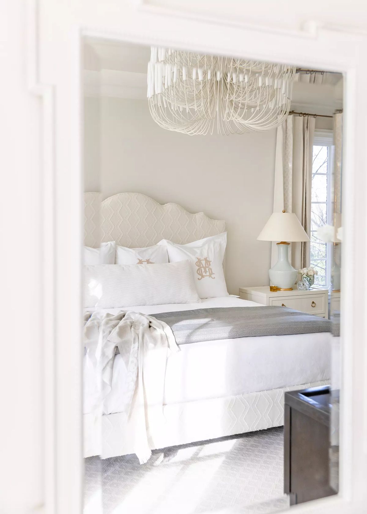 Bedroom with tufted headboard, gray bedspread, nightstands, chandelier, large windows, and a mirrored dresser.