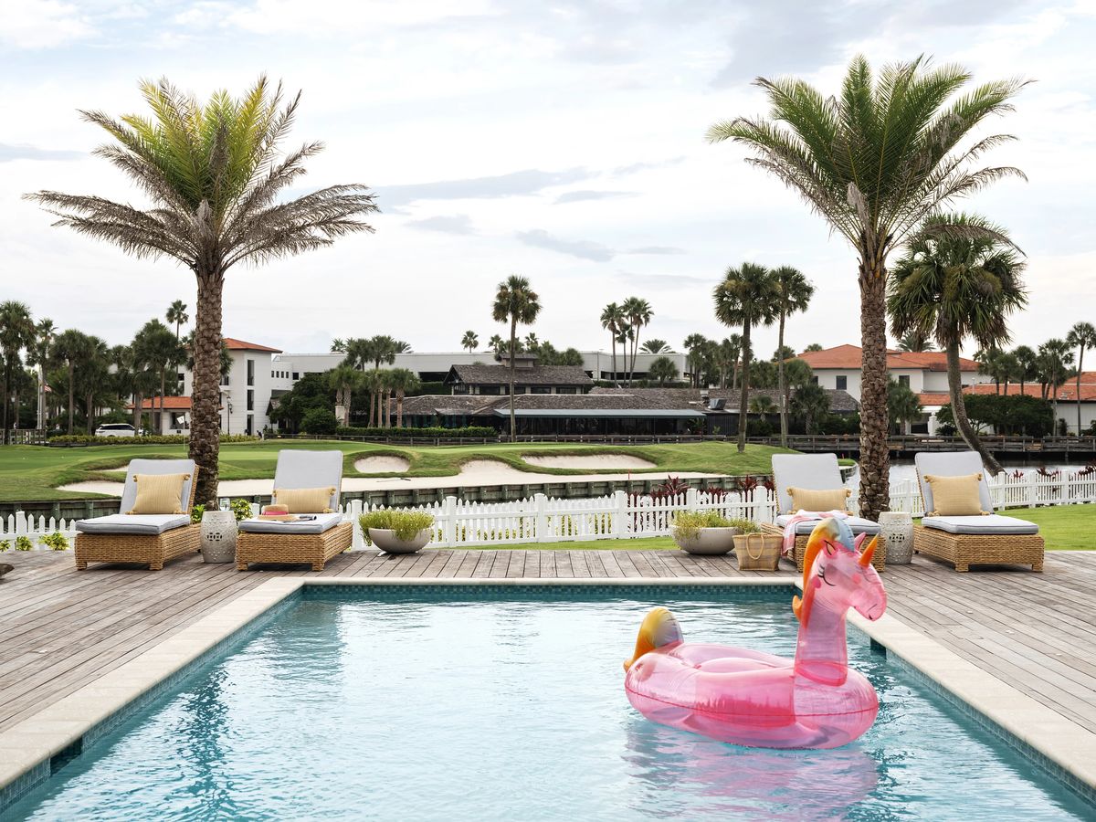 Patio with swimming pool, pink unicorn float, lounge chairs, and palm trees overlooking a golf course.