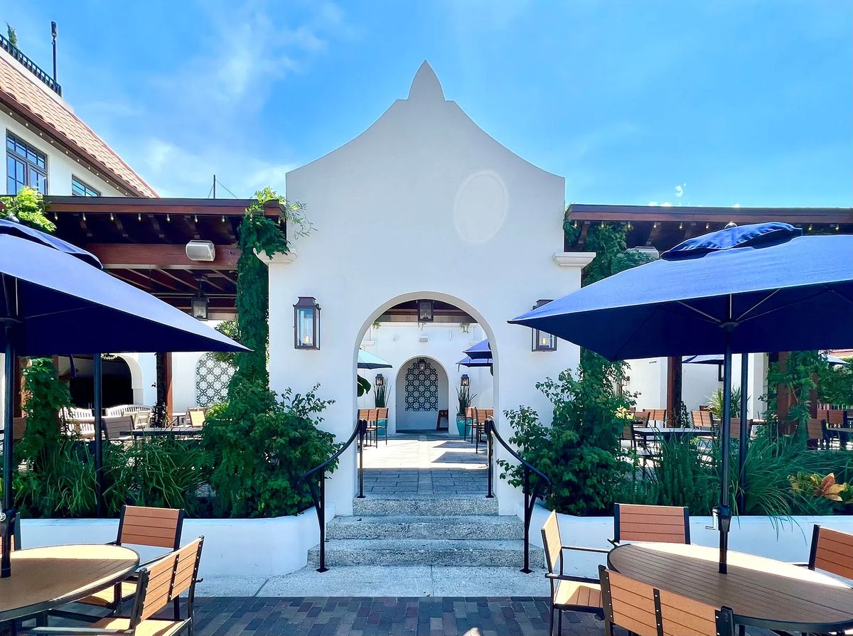 Exterior front area with arched entryway, wooden tables, blue umbrellas, and surrounding greenery.