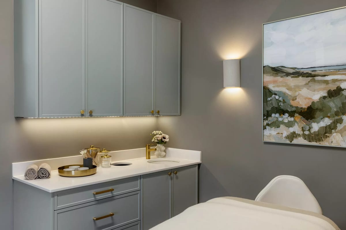 Bathroom with gray cabinetry, white countertop, gold faucet, landscape painting, and towel rack.