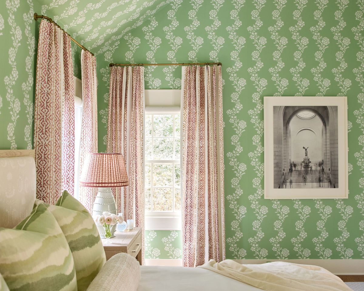 Bedroom with bed, patterned pillows, bedside lamp, green floral wallpaper, and red curtains.