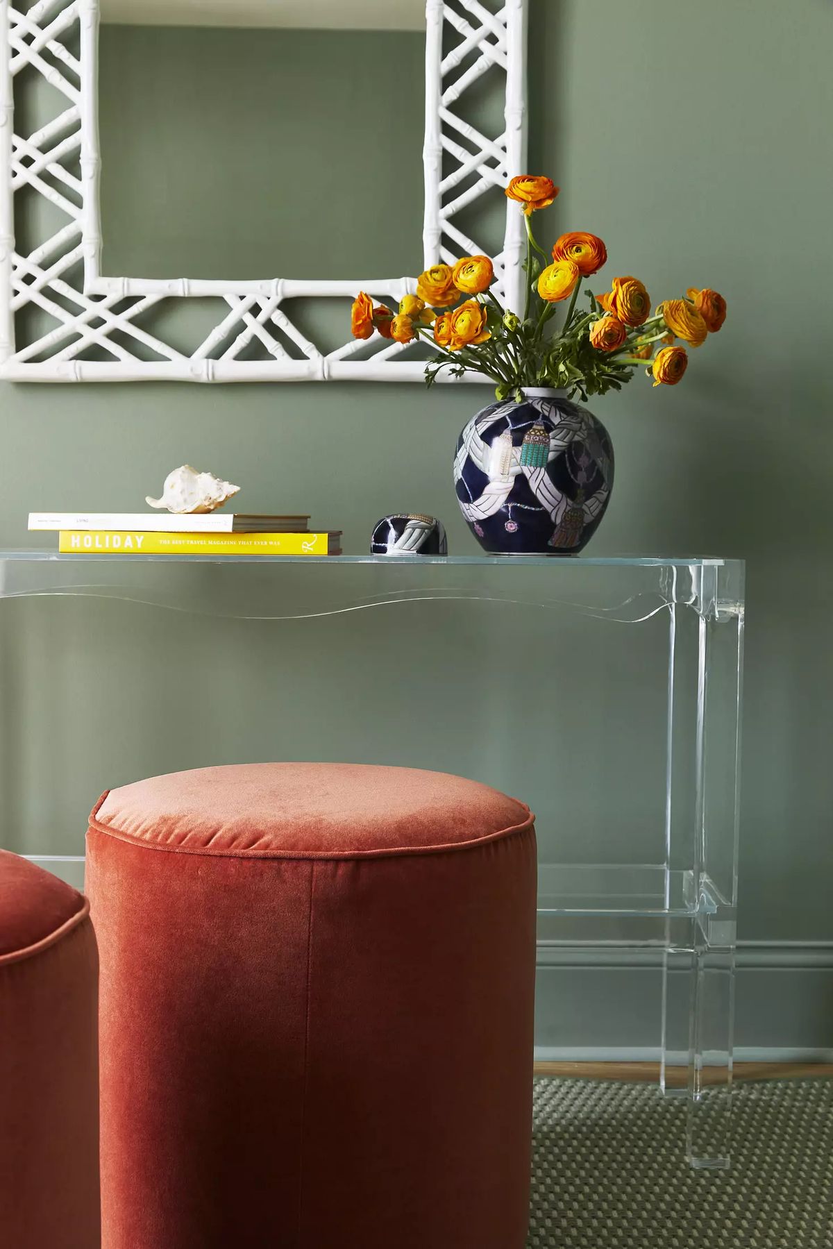 Wall with green color, acrylic console table, orange stools, flowers, book, and decorative mirror.