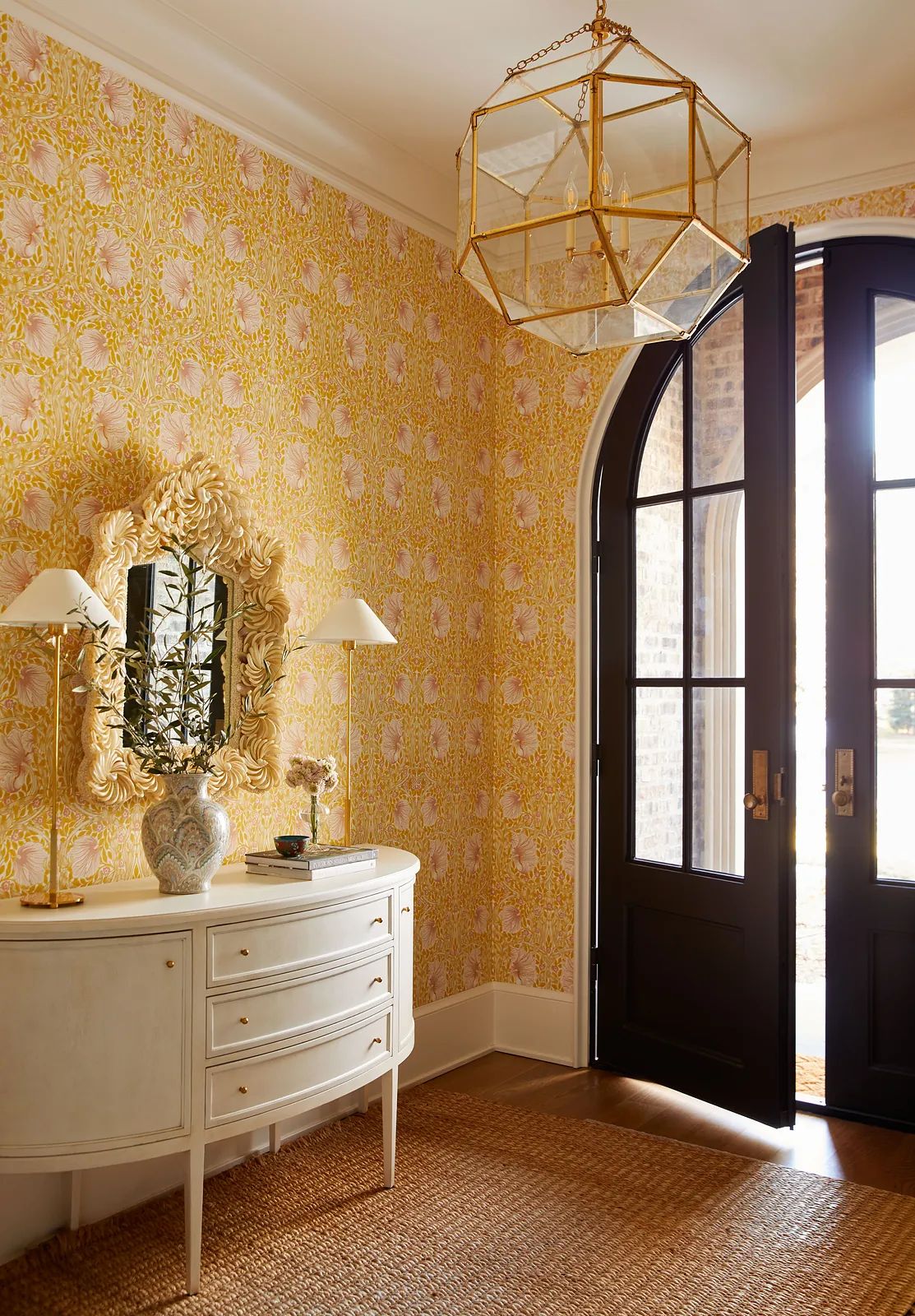 Foyer with white console table, decorative mirror, yellow floral wallpaper, and arched black doors.