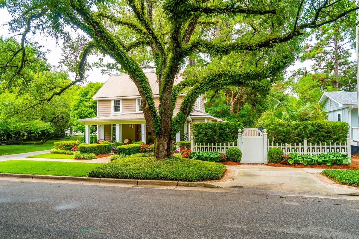 Front exterior of a house with beige siding, metal roof, manicured lawn, white picket fence, and large tree.