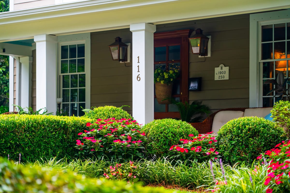 Exterior front with porch, wooden door, lanterns, trimmed bushes, and colorful flower beds.