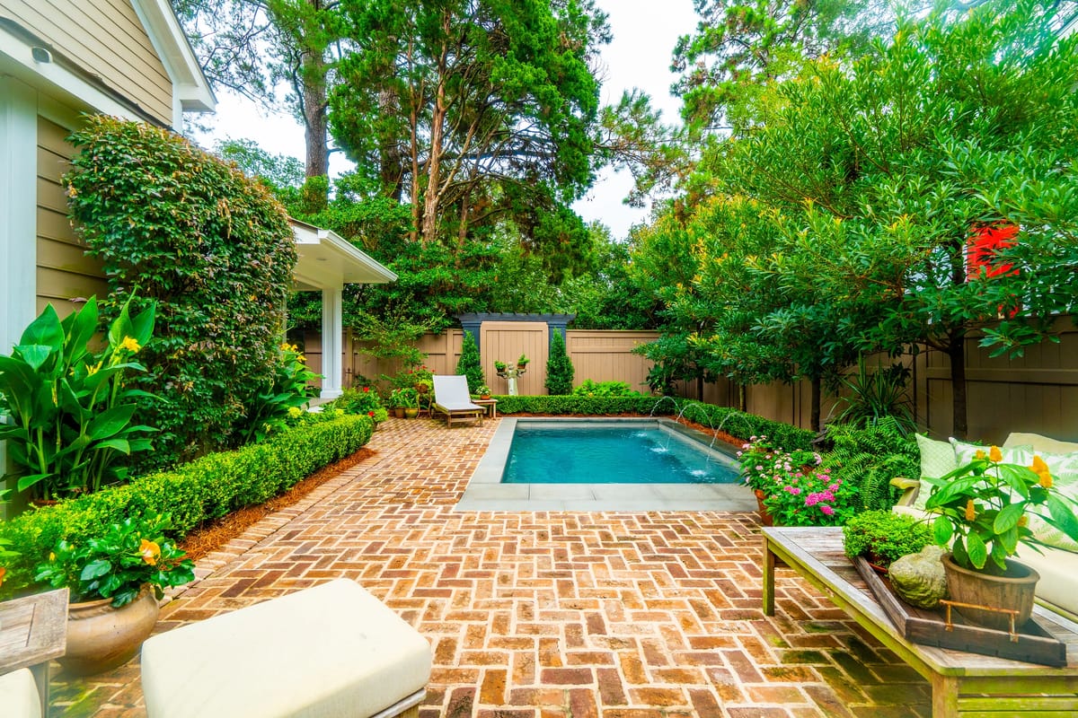 Garden with rectangular pool, brick patio, shrubs, flowers, and a lounge chair.