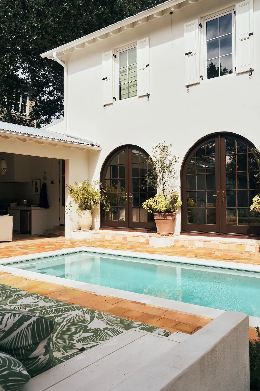 Exterior rear with small pool, tiled deck, arched doors, and potted plants against white walls and green shutters.