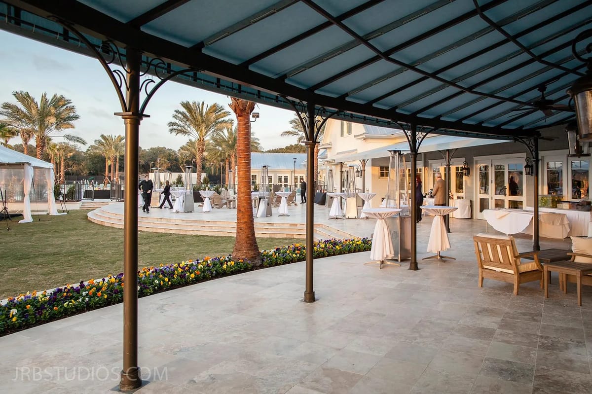 Patio with stone floor, wooden chairs, high-top tables, blue pergola, and palm trees in the background.
