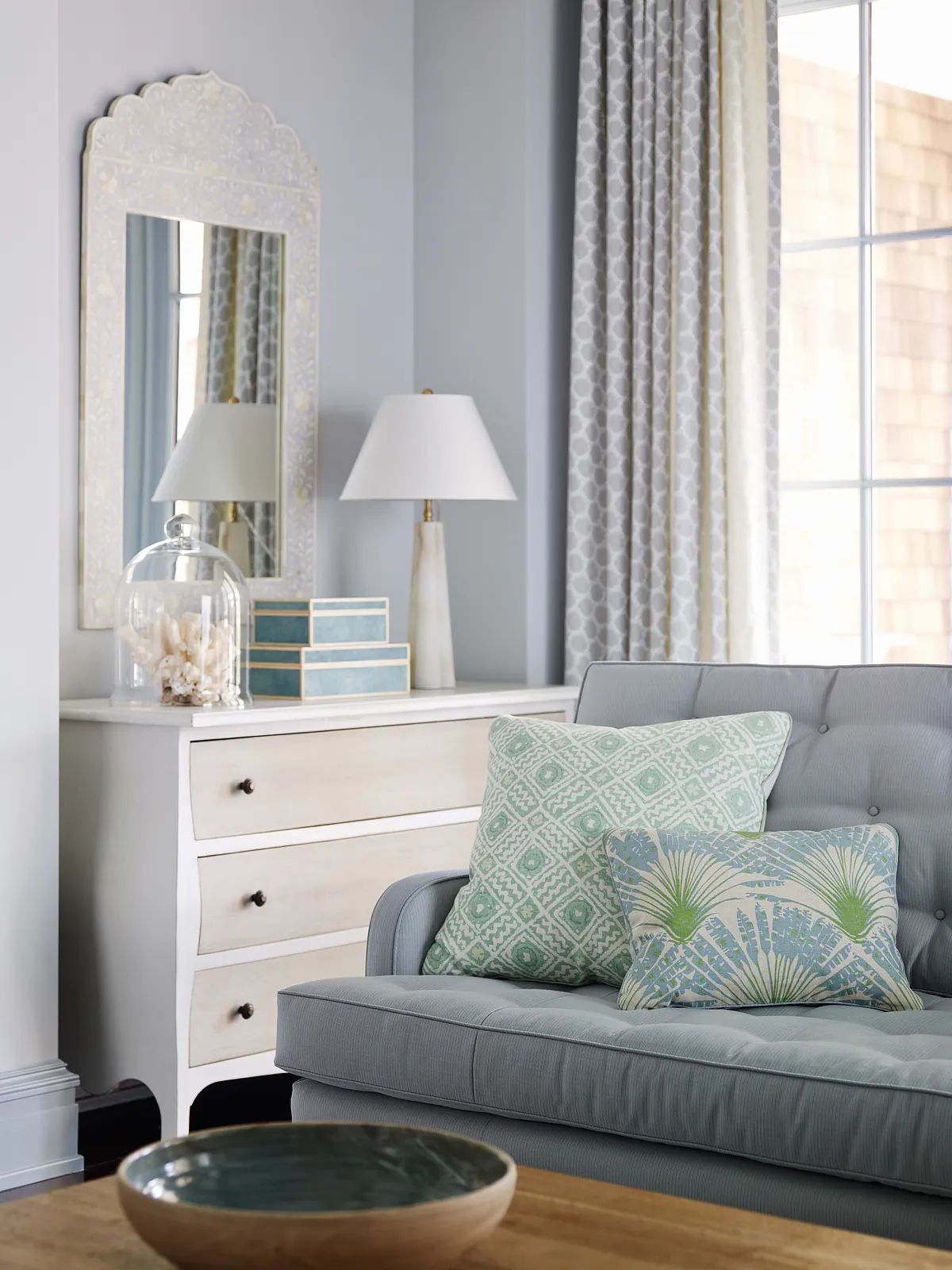Living room with gray sofa, decorative items, dresser, and large windows with patterned curtains.