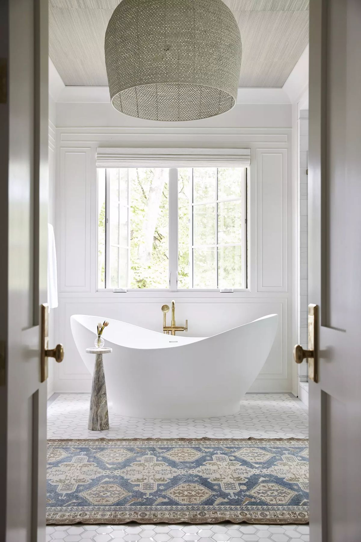 Bathroom with freestanding white bathtub, marble table, area rug, and large windows with gold fixtures.