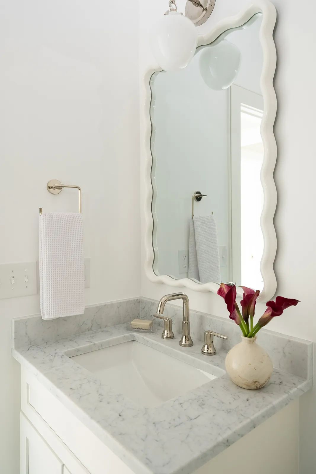Bathroom with white vanity, marble countertop, scalloped mirror, chrome faucet, and calla lilies.