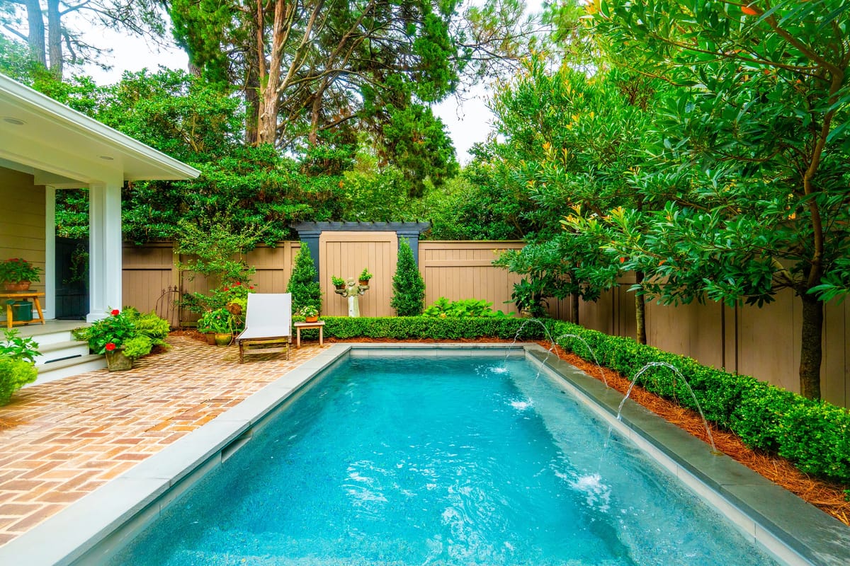 Outdoor garden with swimming pool, brick tiles, lounge chair, and surrounding greenery.