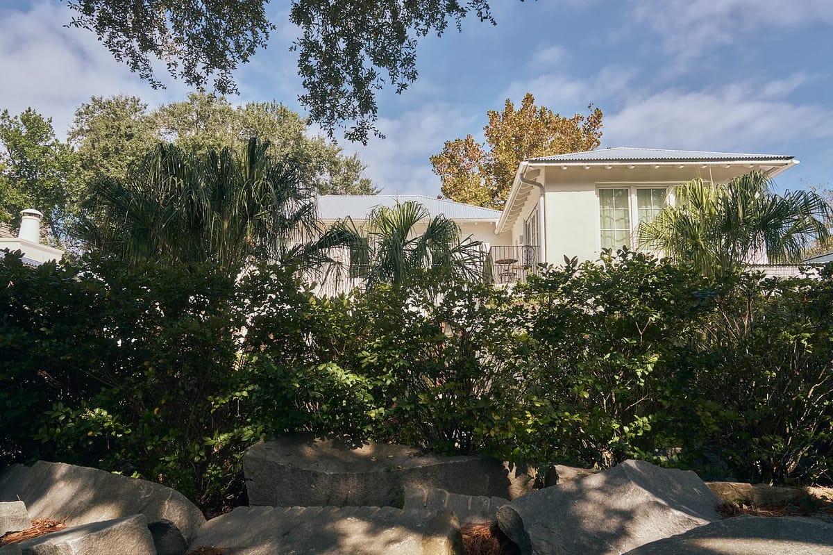 Front exterior of a house with white facade, windows, balcony, surrounded by palm trees and rocks.