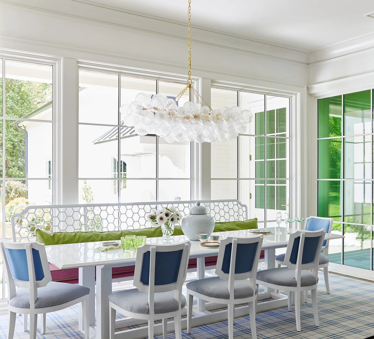 Dining room with white table, blue and white chairs, light fixture, windows, sideboard, and floral decor.