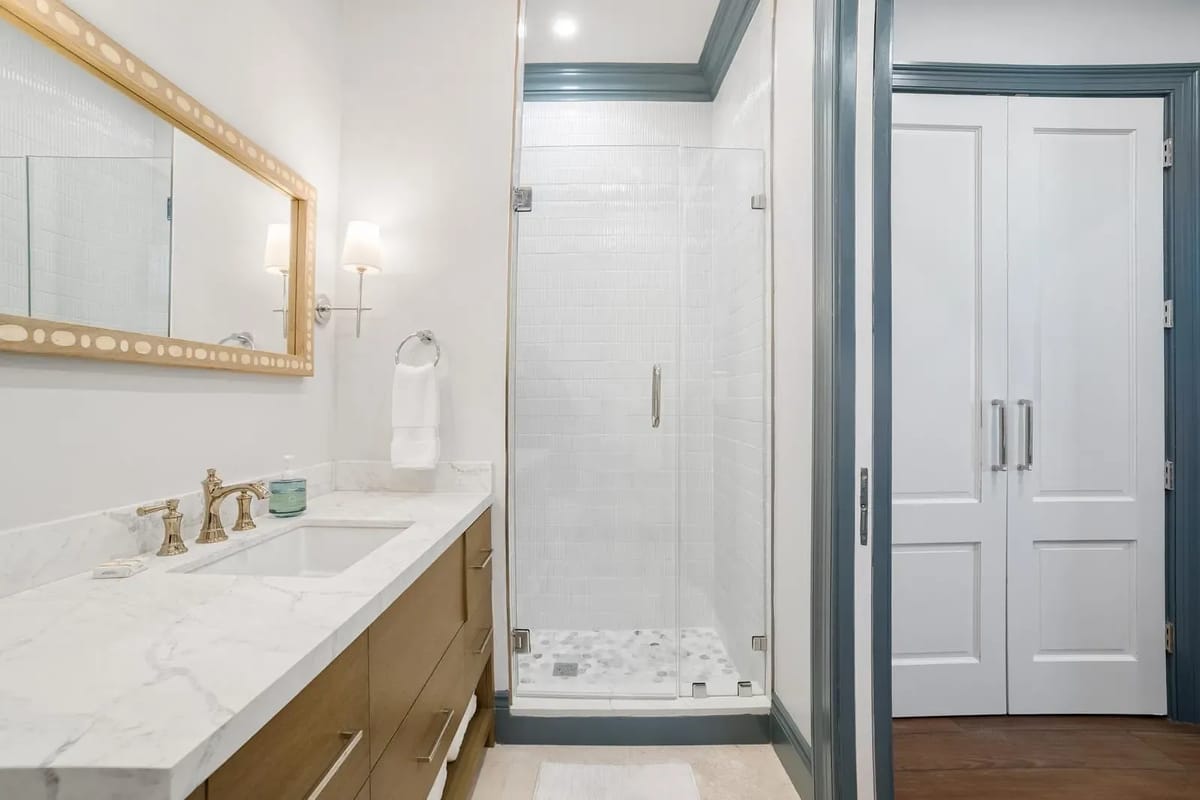 Bathroom with double vanity, marble countertop, gold faucets, glass shower enclosure, and white walls with blue trim