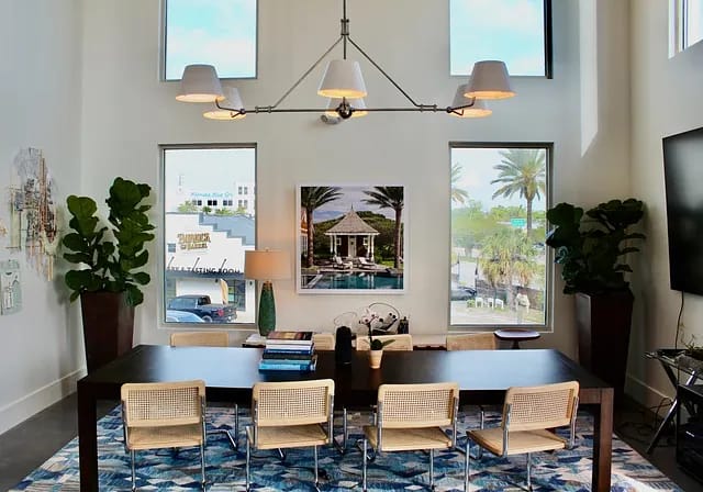 Dining room with large wood table, eight woven chairs, chandelier, two tall windows, and wall painting.