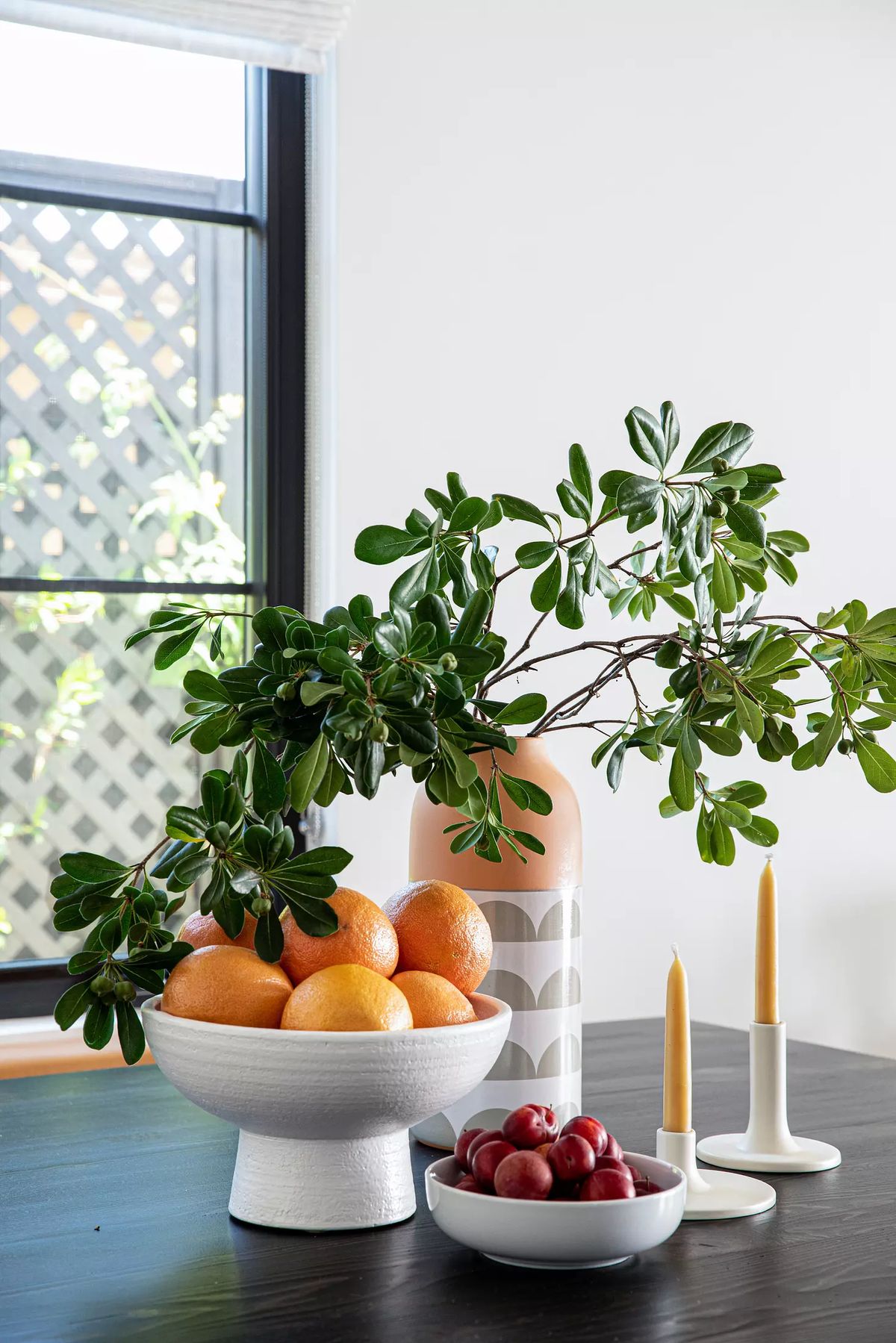 Decorative arrangement with fruits, a vase, and a dark tabletop near a window with lattice pattern.