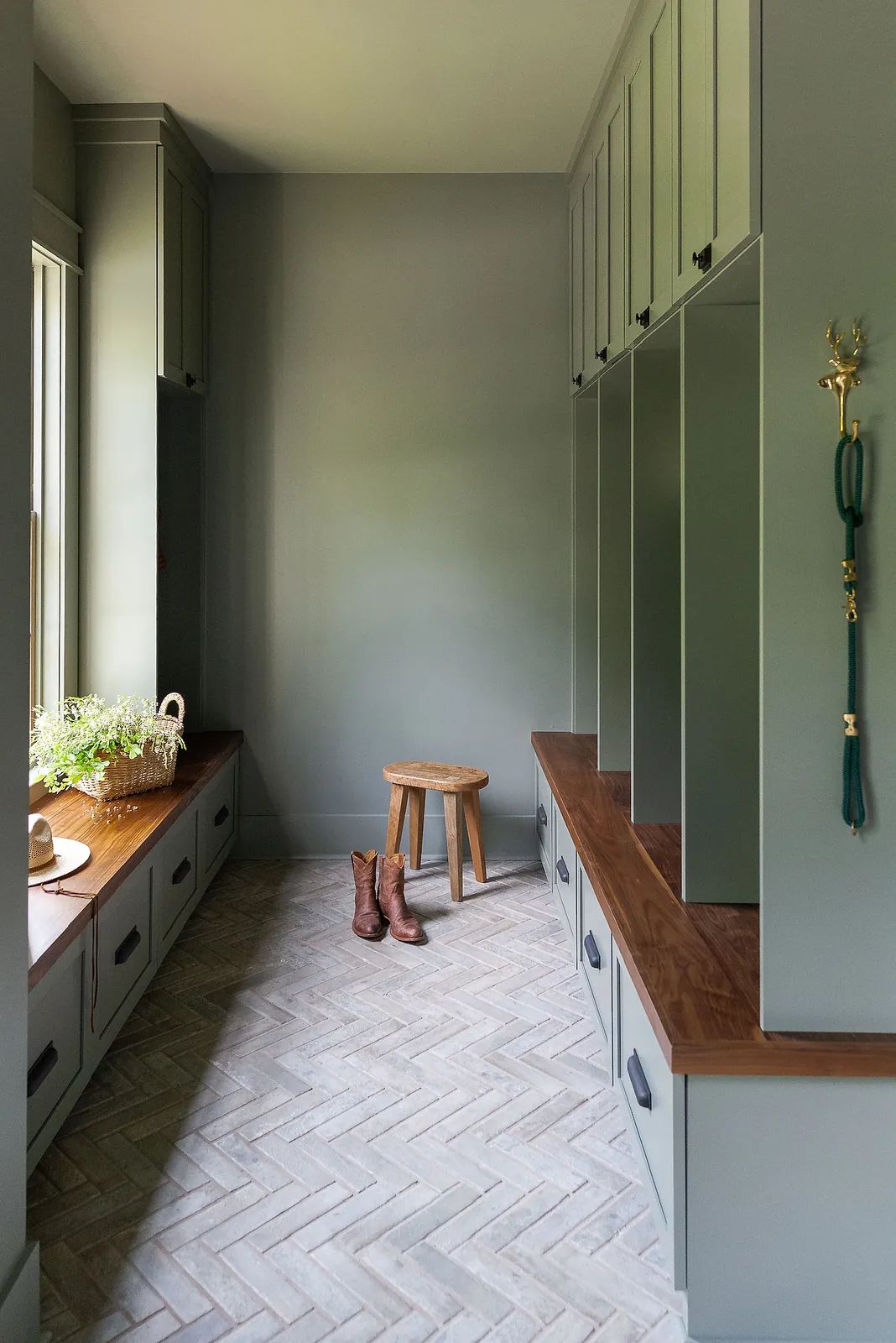 Mudroom with green cabinets, wooden bench, herringbone flooring, stool, boots, and plants in a basket