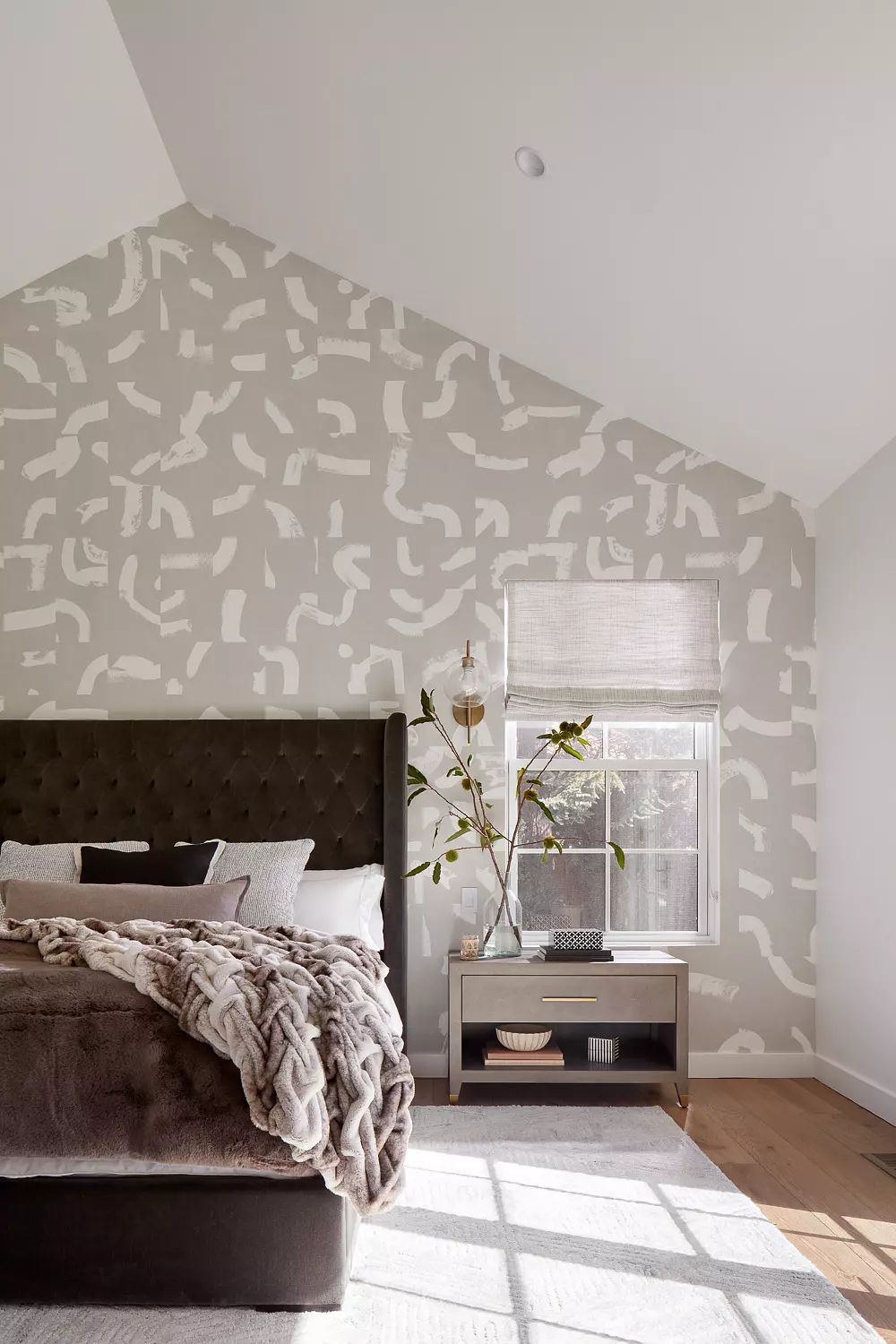 Bedroom with tufted bed, gray blanket, pillows, side table, patterned wall, and wooden floor.