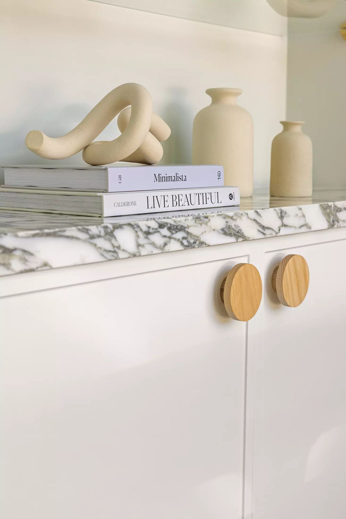 Display of white cabinet with marble top, beige vases, book, and sculptural object.