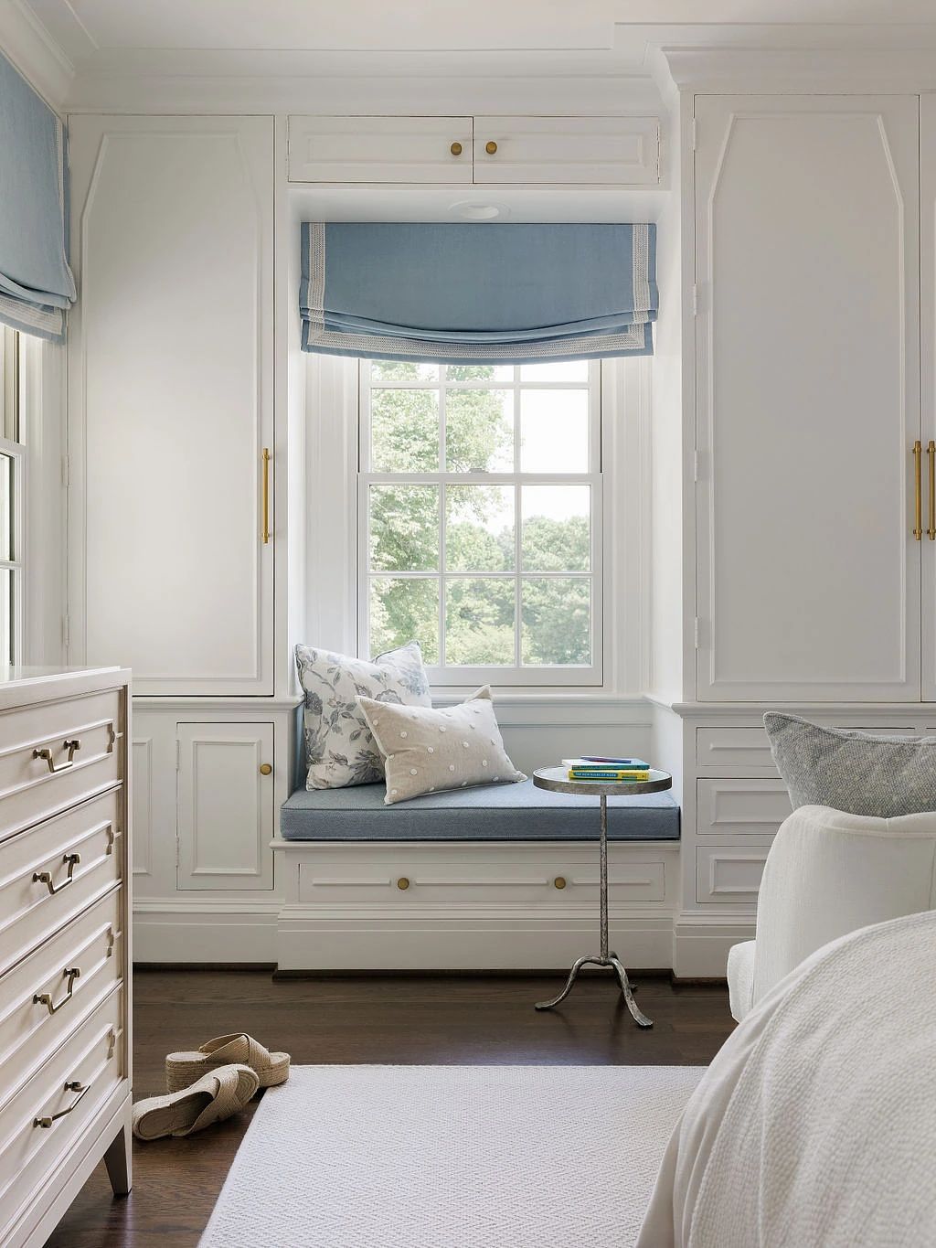 Bedroom with window seat, white dresser, side table, and blue window shades.