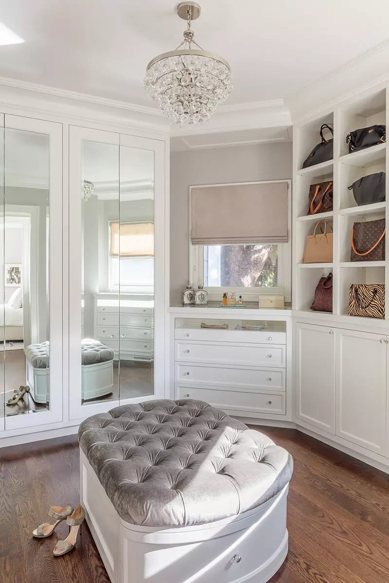 Closet with tufted ottoman, mirrored wardrobe, shelves with handbags, and hardwood flooring.