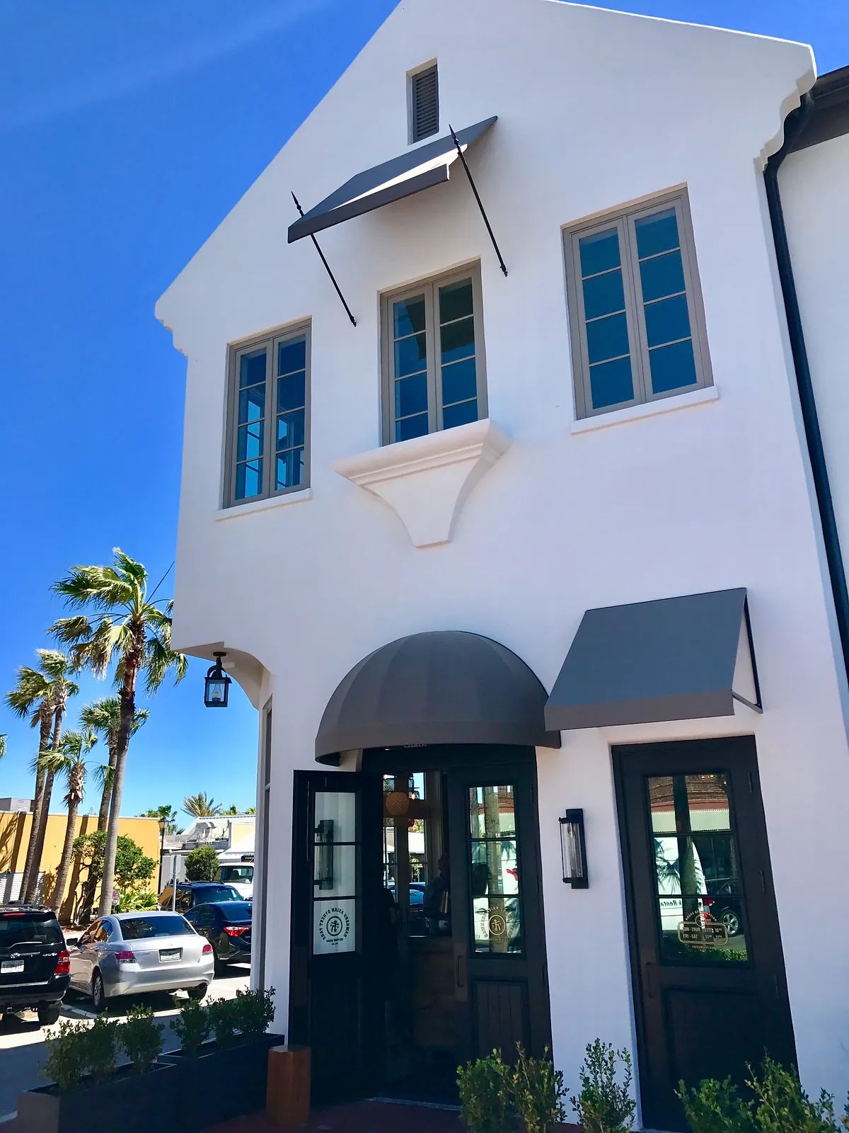 Exterior front featuring a white building with arched entry, black doors, gray and black awnings, and blue windows.