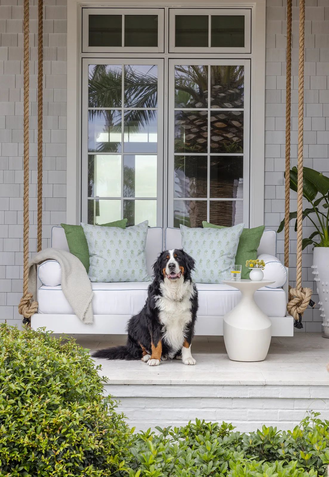 Patio with white sofa, green cushions, side table, swing, and greenery.