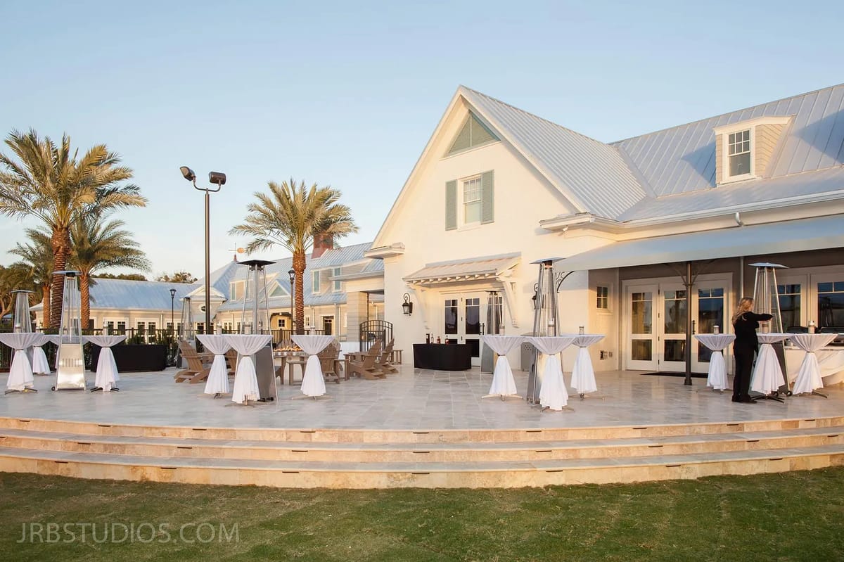 Exterior space with round tables, palm trees, stone flooring, and a bar setup in front of a white building.