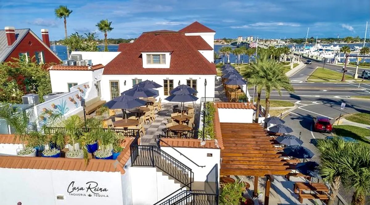 Exterior front view with red-tiled roof, outdoor dining area, umbrellas, palm trees, and decorative wall.
