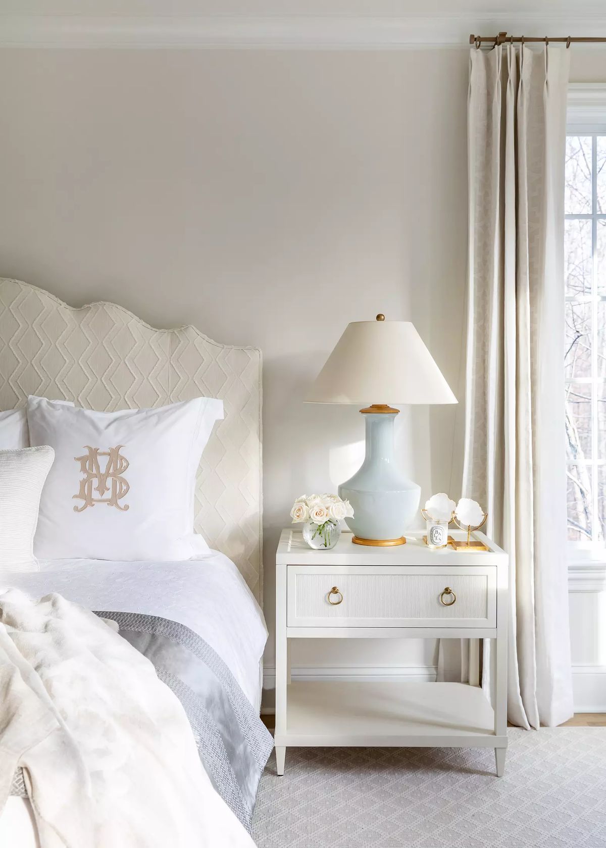 Bedroom with patterned headboard, white bedding, nightstand, lamp, and large window with white curtains