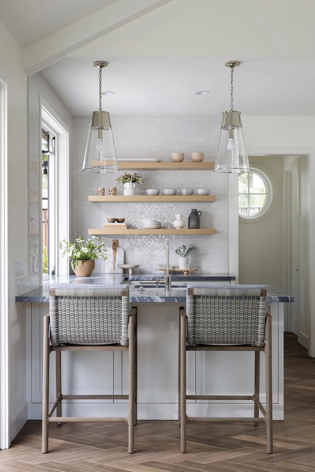 Kitchen with bar, high stools, wooden shelving, sink, and natural light from windows.