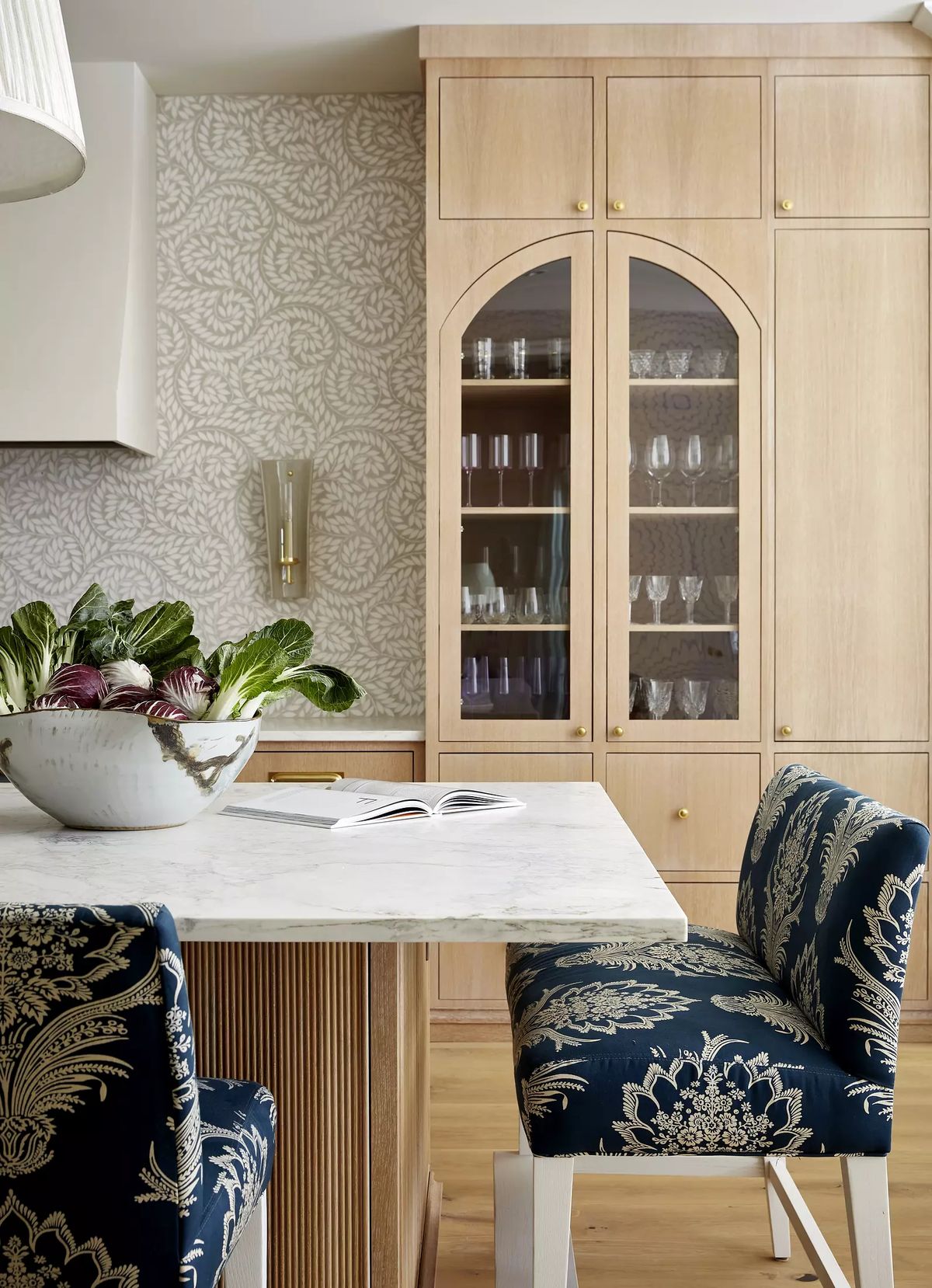 Kitchen with marble island, light wood cabinetry, glass-fronted cabinet, and floral upholstered chairs.