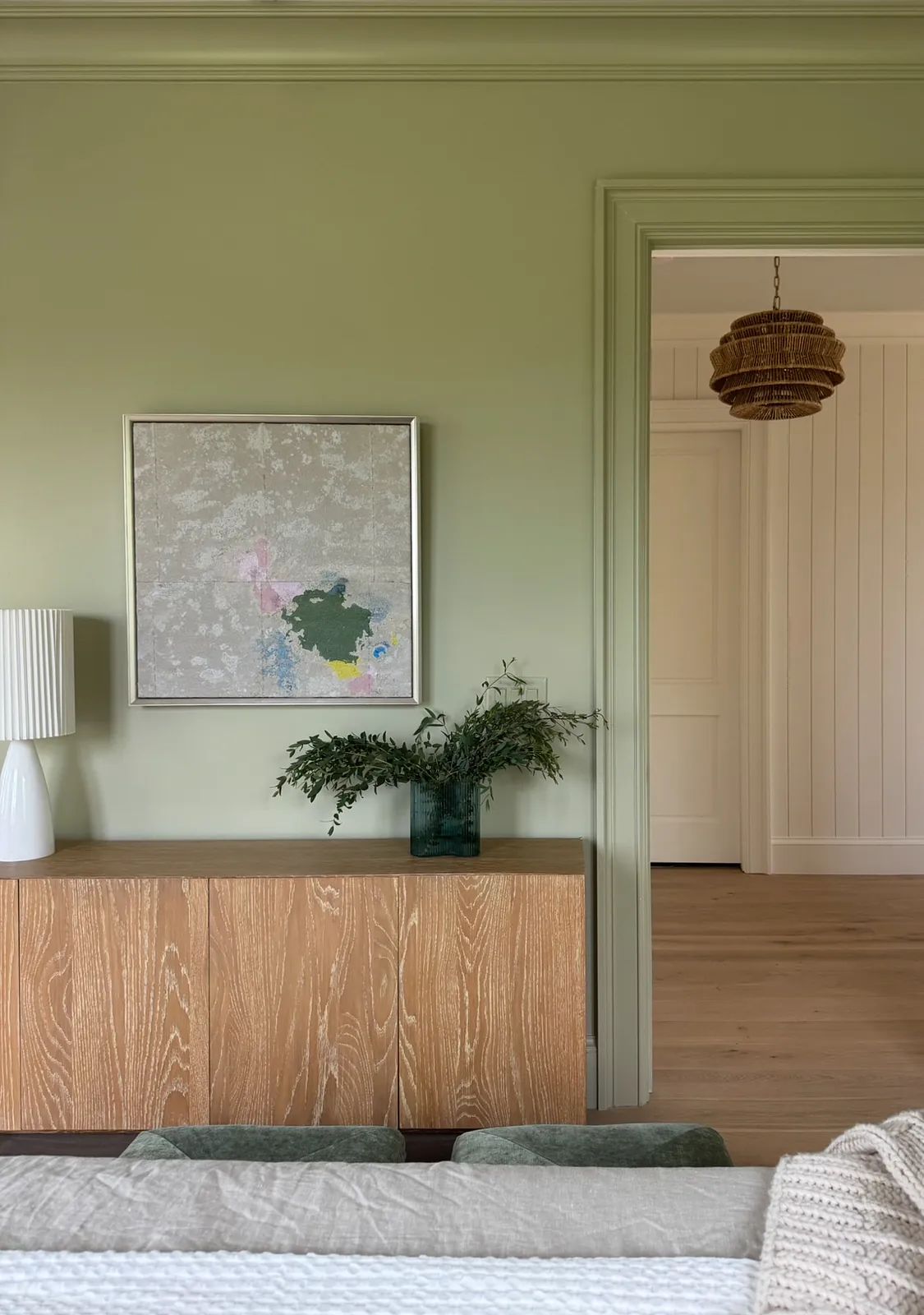 Living room with light green walls, wooden sideboard, artwork, foliage vase, and a white lamp.