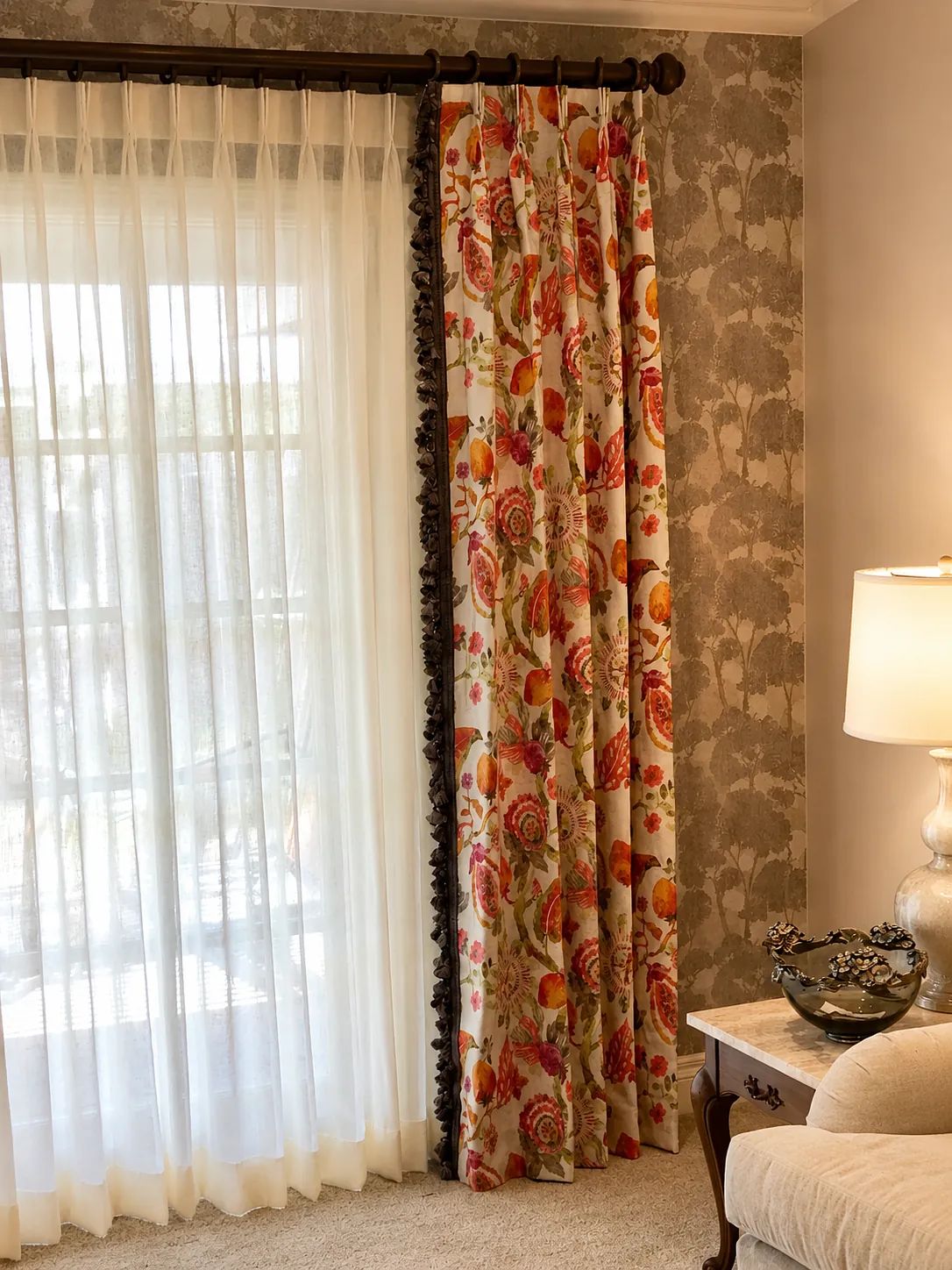 Living room corner with floral curtain, sheer drapes, side table, lamp, and patterned wallpaper.