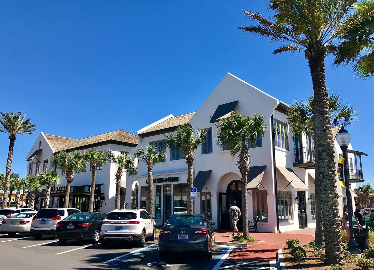 Exterior view of shopping complex with palm trees, white buildings, and parked cars.