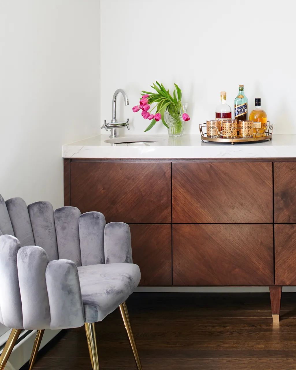 Butler's pantry with dark wood cabinet, white countertop, sink, grey chair, bottles, and vase with tulips