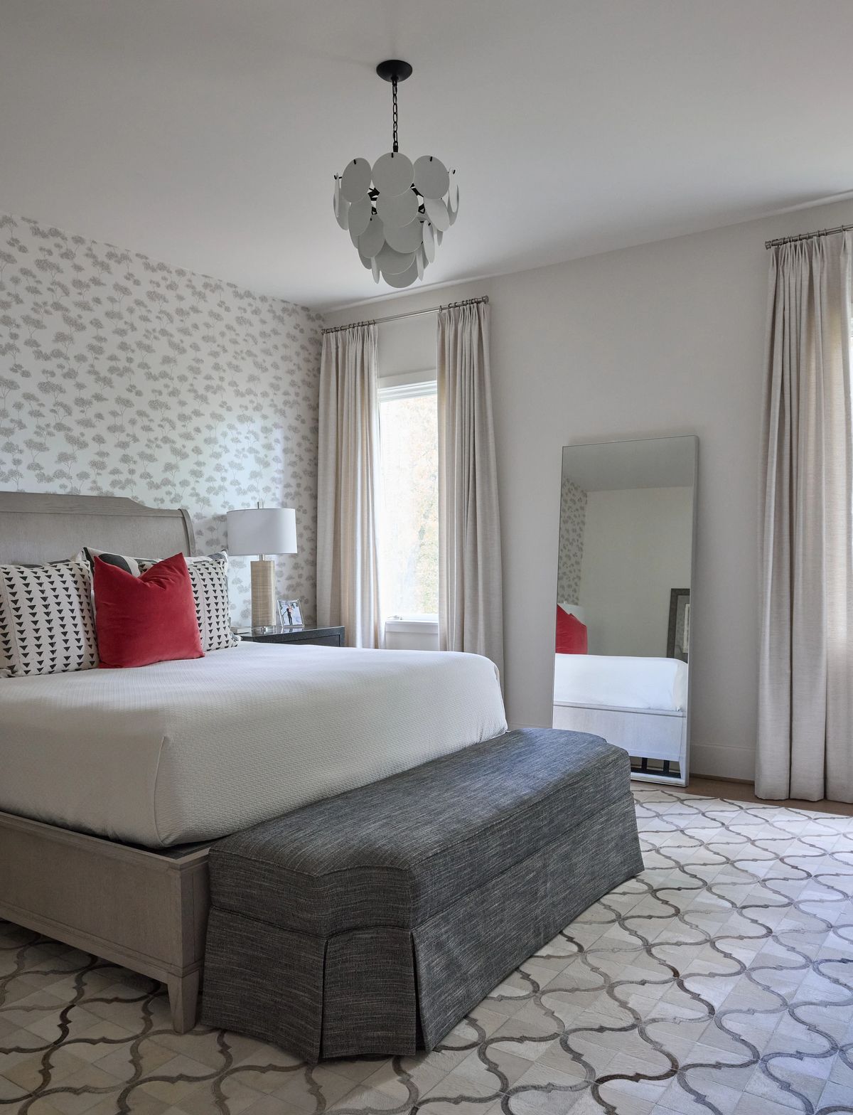 Bedroom with gray upholstered bed, red accent pillow, nightstand, lamp, mirror, and patterned wallpaper