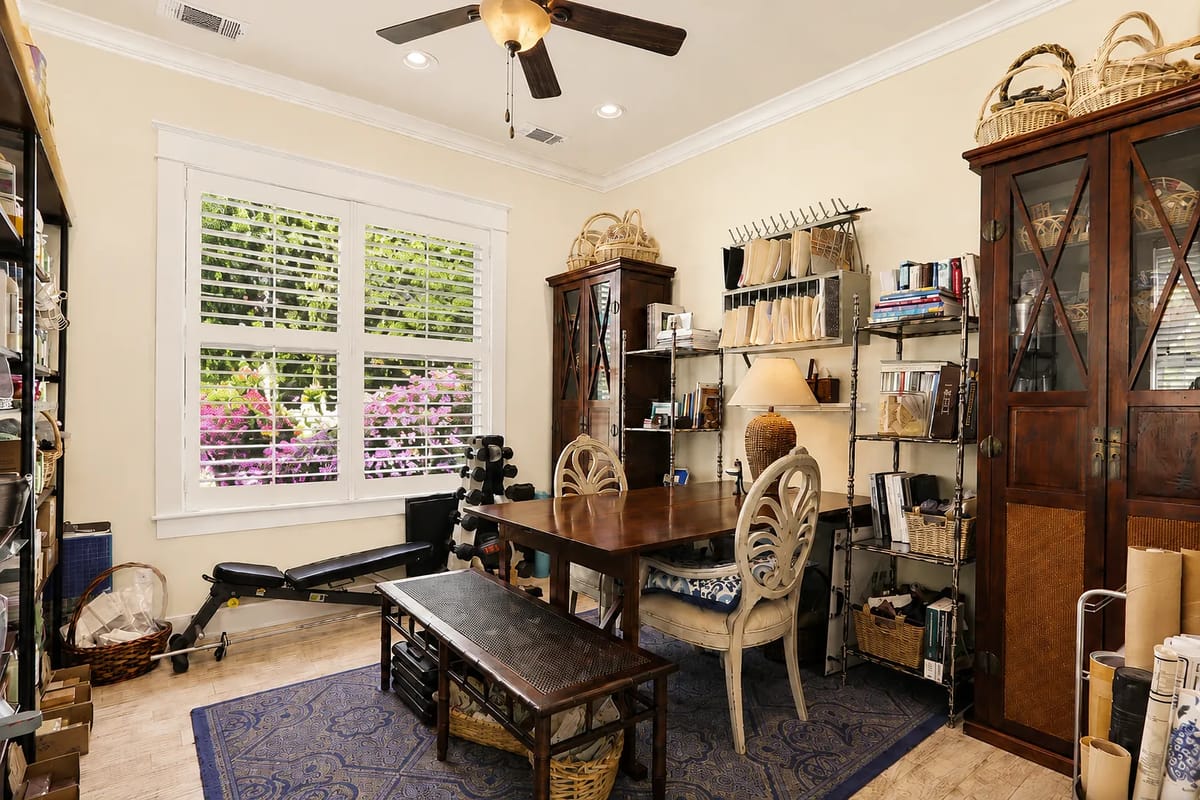 Office with wooden table, decorative chairs, shelves, rowing machine, and large window with plantation shutters.