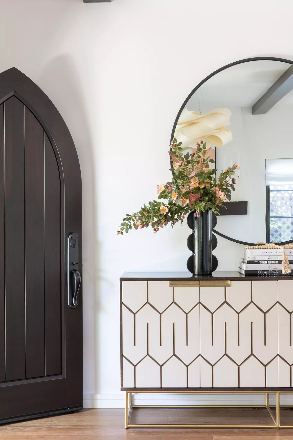 Foyer with dark wood door, geometric sideboard, round mirror, vase with flowers, and stack of books.