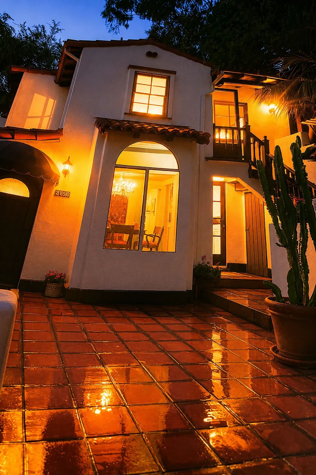 Exterior view of a two-story home with terracotta tiles, glowing windows, and a tall cactus.