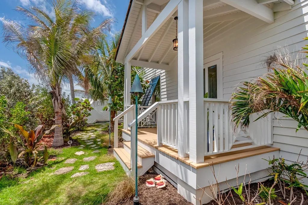 Exterior front view with covered porch, steps, white railings, palm trees, and tropical plants.
