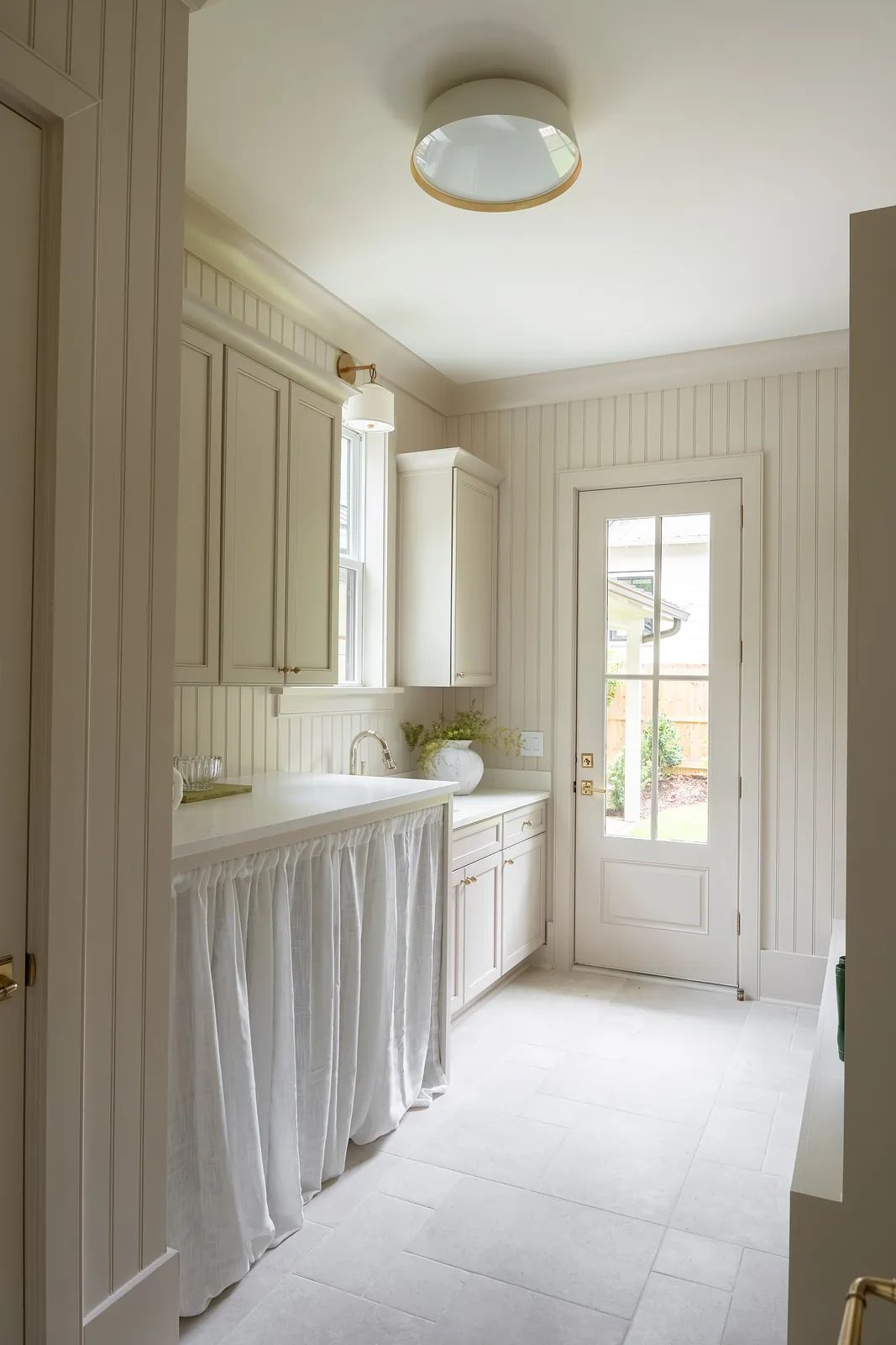 Laundry room with white cabinetry, fabric skirted countertop, beige tiled floor, and door leading outside.