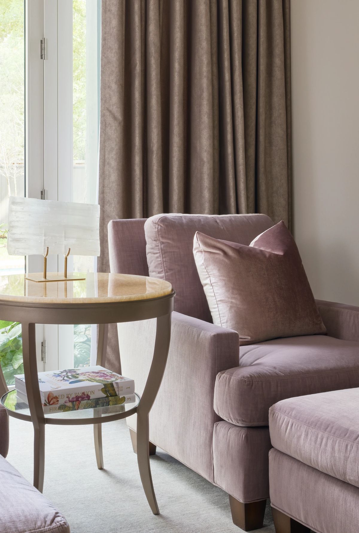 Living room with pink armchairs, glass side table, stack of books, large windows, and brown curtains.