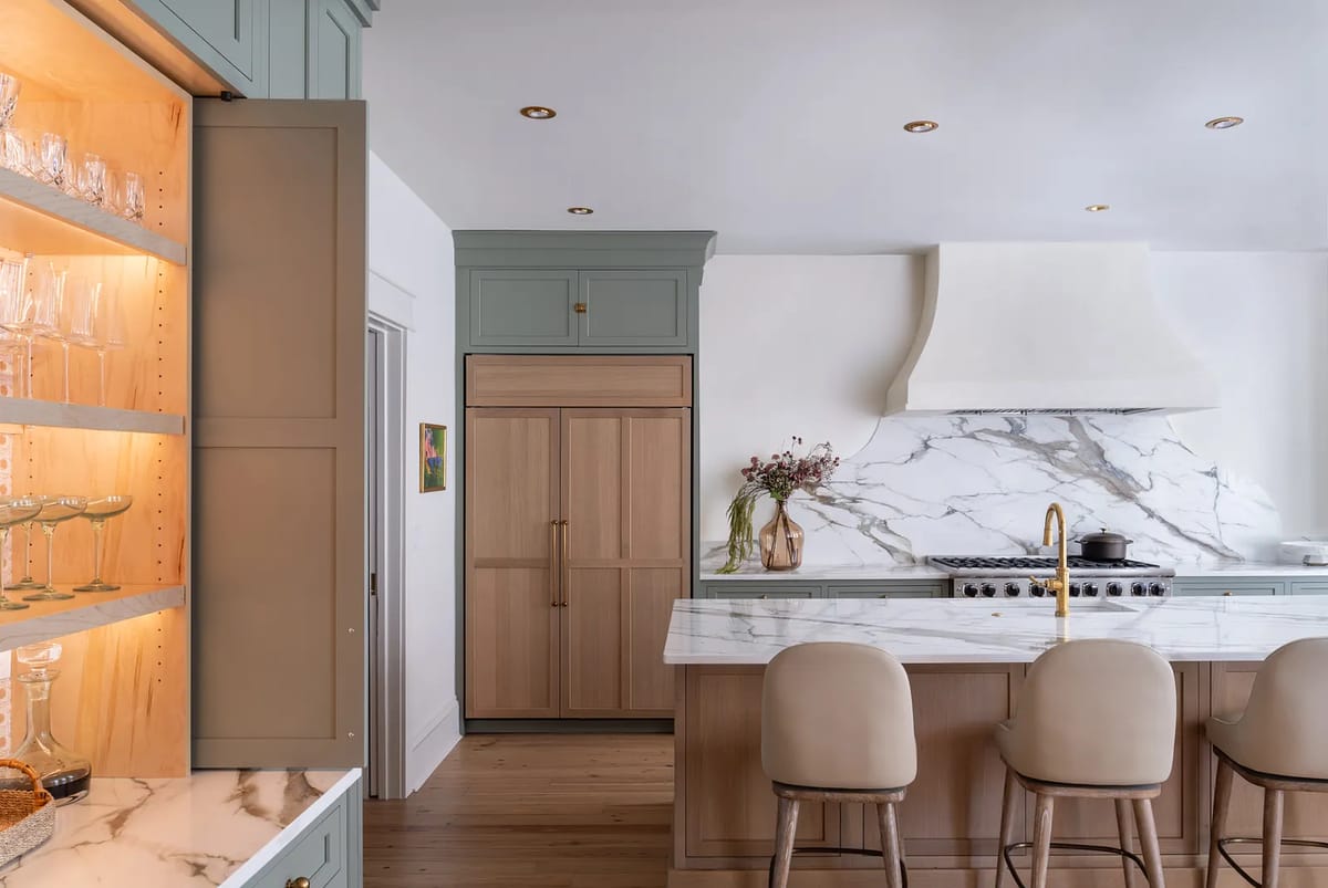 Kitchen with light green cabinetry, wood refrigerator, marble countertop, beige bar stools, and glass display cabinet.