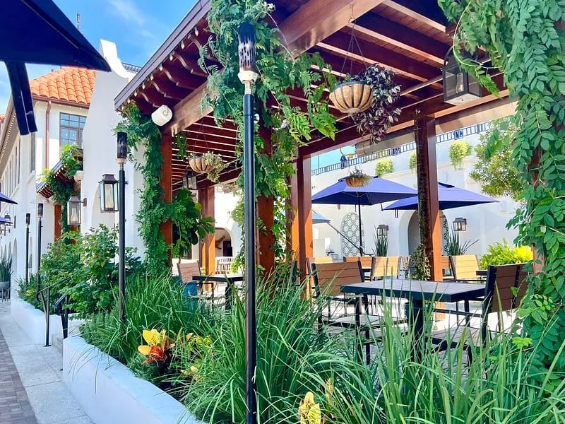 Patio with wooden pergola, outdoor tables, chairs, umbrellas, and surrounding greenery.
