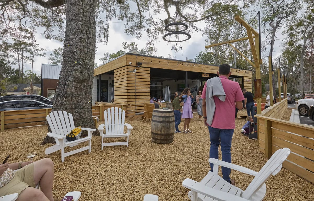 Outdoor patio with white adirondack chairs, wooden barrel table, and rustic structure surrounded by trees.