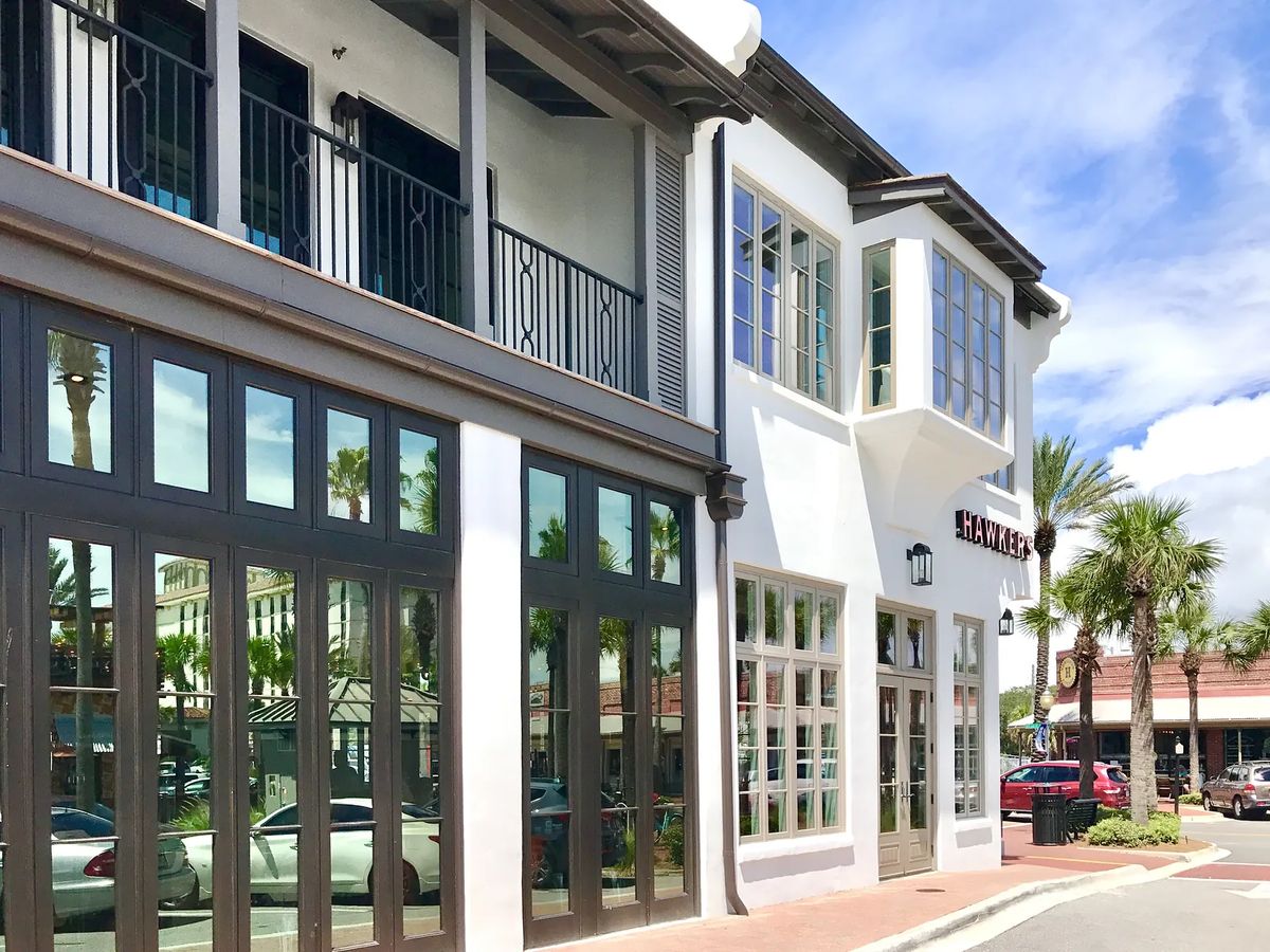 Modern commercial exterior with glass windows, balcony, potted palms, and brick pavement.